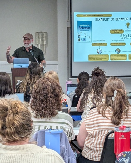 Presenter at a lectern delivering behavior-science training to educators, with a projected slide on the hierarchy of behavior in education