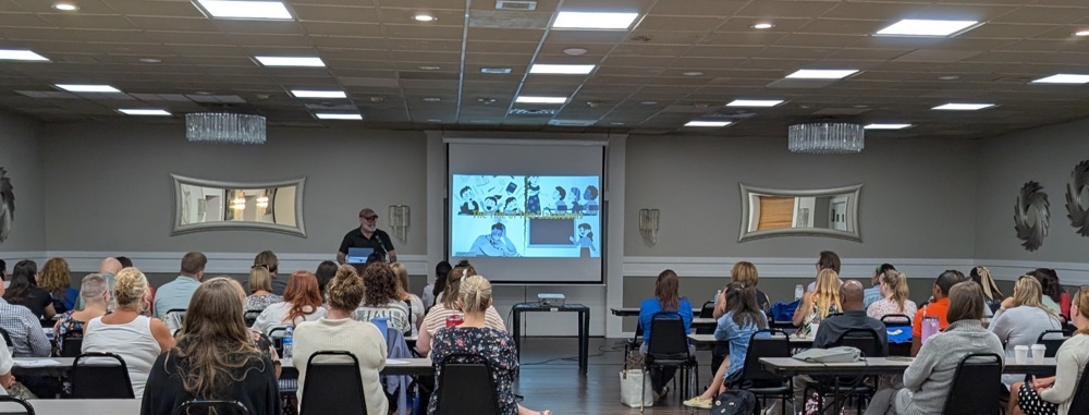 Dr. Gavoni presenting to a large room of educators seated at tables, beside a projected slide titled "The Tale of Two Classrooms"