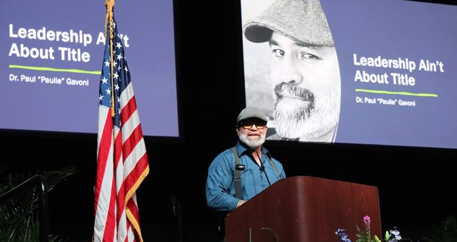 Dr. Gavoni delivering a keynote on a large conference stage beside a U.S. flag