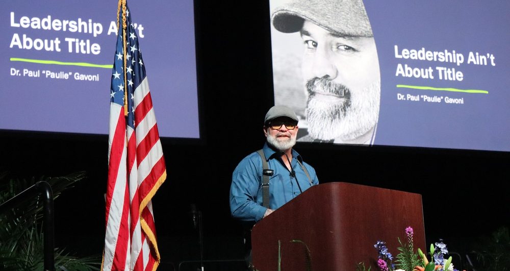 Dr. Paul "Paulie" Gavoni delivering the keynote "Leadership Ain't About Title" on a large conference stage in front of a U.S. flag